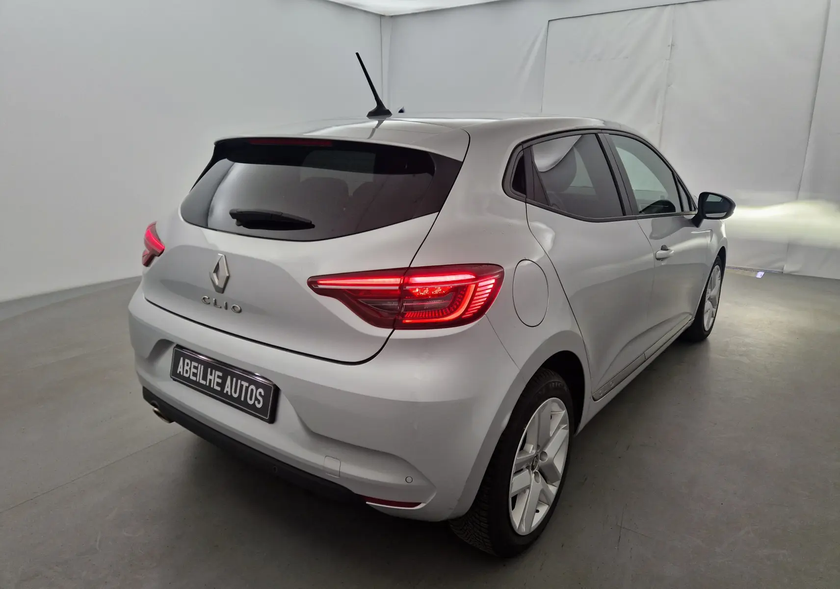 Vue 3/4 arrière droite d'une Renault Clio V gris platine avec feux arrière allumés dans un studio blanc.