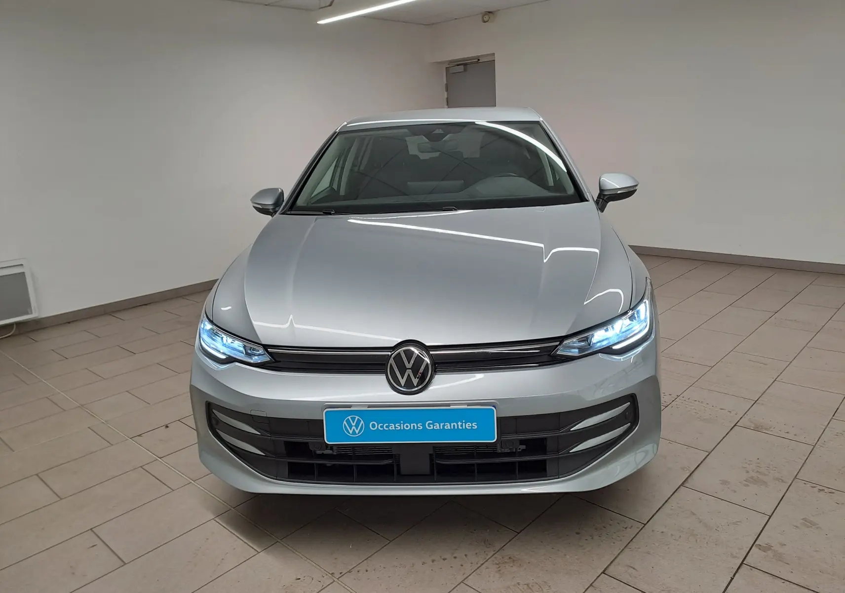 Vue avant d'une Volkswagen Golf 1.5 eTSI 2025 en Argent Dolomite avec phares LED allumés dans un intérieur neutre.