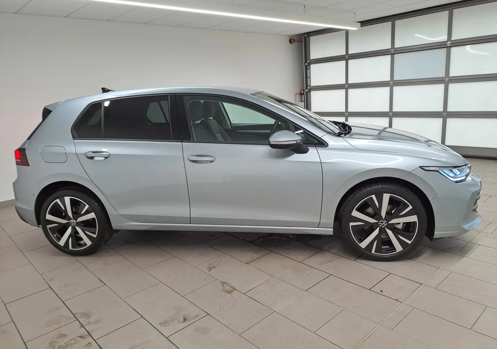 Profil droit d'une Volkswagen Golf 1.5 eTSI 2025 en Argent Dolomite avec jantes alliage 18'' Catania dans un garage.