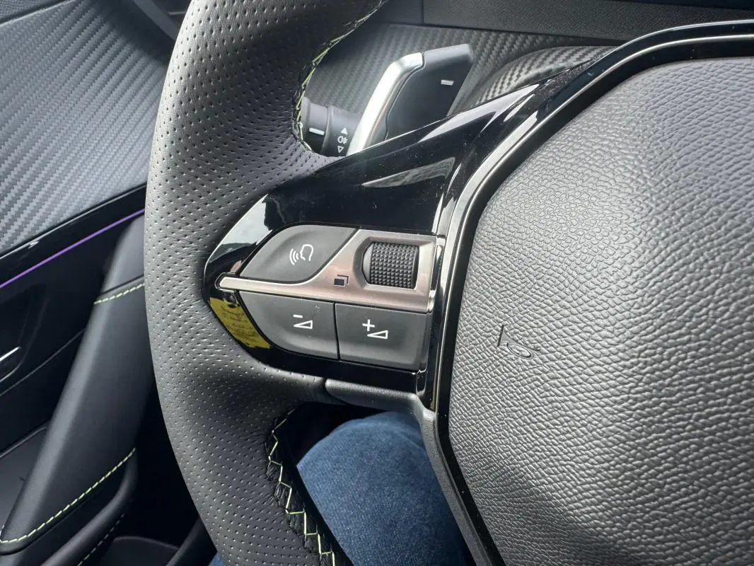 Gros plan sur le côté gauche du volant perforé du Peugeot 208 Hybrid 110 gris, avec commandes audio et finition noire brillante.