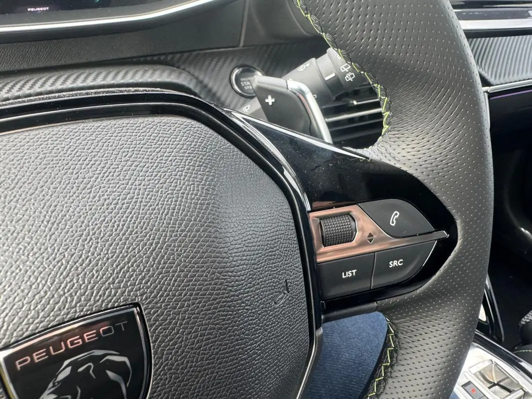 Gros plan sur le volant perforé avec surpiqûres vertes du Peugeot 208 Hybrid 110 e-DCS6 GT, montrant les commandes audio et téléphone.