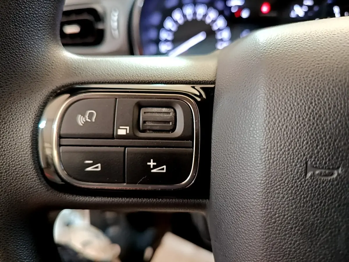 Gros plan sur les commandes de volume et téléphone du volant noir de la Citroën C3 blanche, tableau de bord flou en arrière-plan.