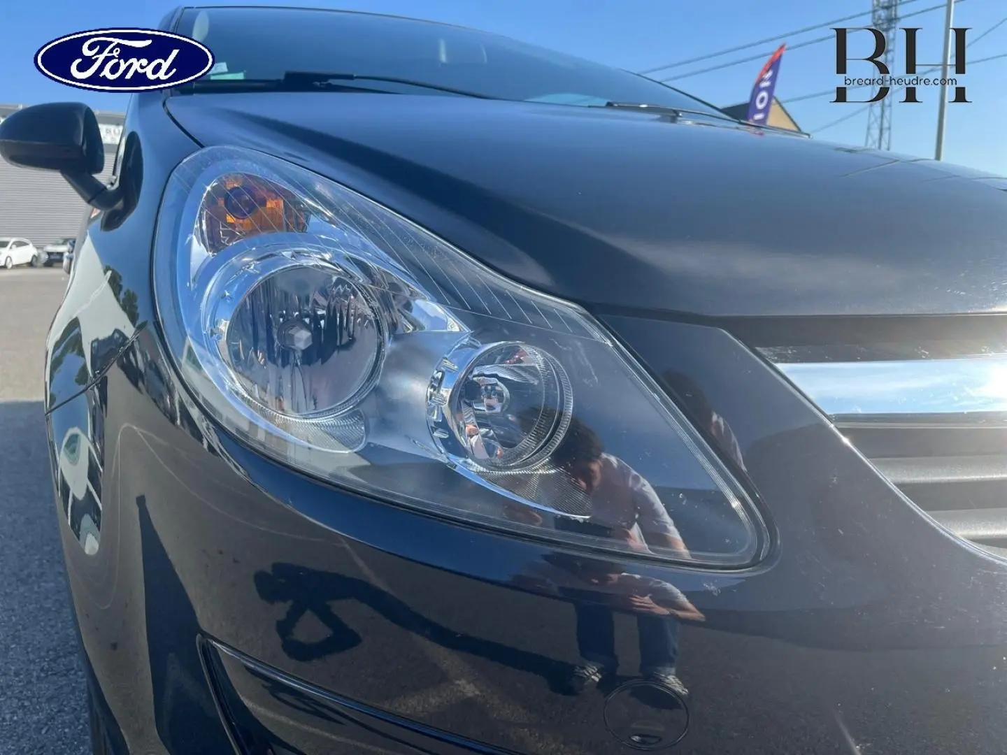 Gros plan sur le phare avant gauche d’une Opel Corsa noire de 2010, mettant en valeur la carrosserie brillante.