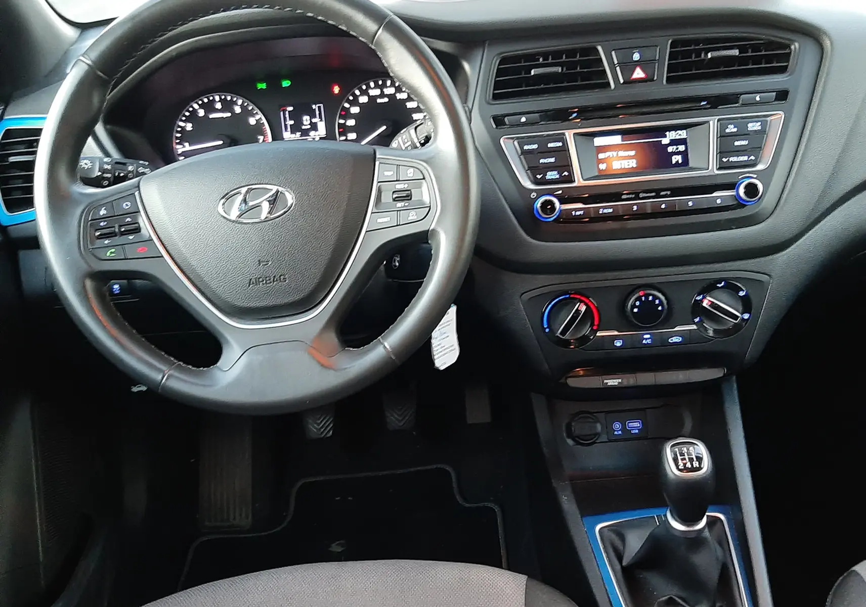 Vue intérieure de face du poste de conduite d'une Hyundai i20 blanche, volant multifonction et levier de vitesse manuel visibles.