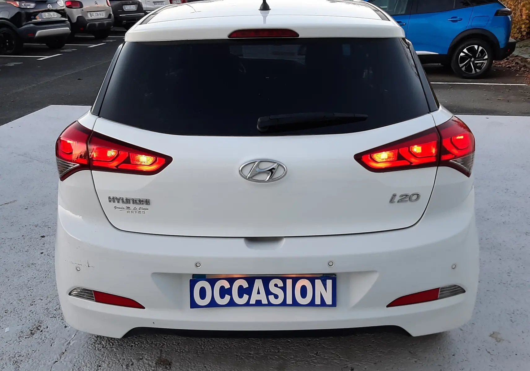 Vue arrière d'une Hyundai i20 blanche avec feux allumés et plaque "OCCASION" sur un parking extérieur.