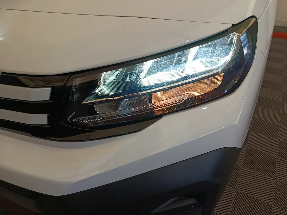 Gros plan sur le phare avant droit du Peugeot Partner blanc 2025, avec détails du contour noir et clignotant orange.