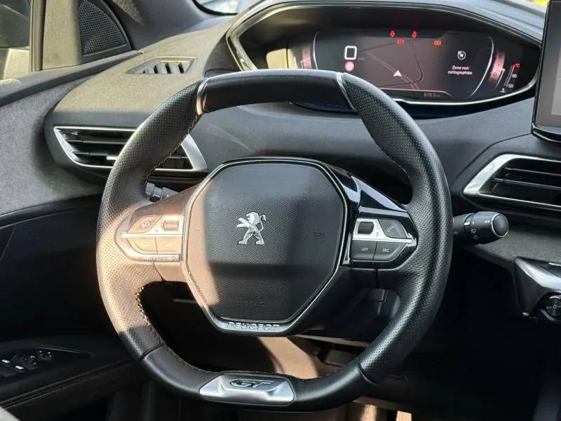 Vue rapprochée du volant multifonction du Peugeot 3008 2024 avec logo lion et finition GT, tableau de bord numérique en arrière-plan.