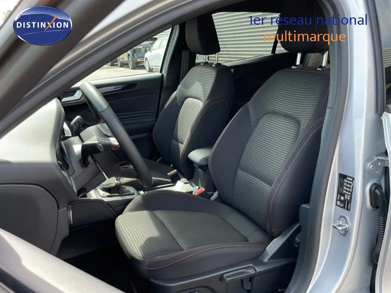 Vue intérieure côté conducteur de la Ford Focus SW gris lunaire, sièges tissu noir avec surpiqûres rouges visibles.