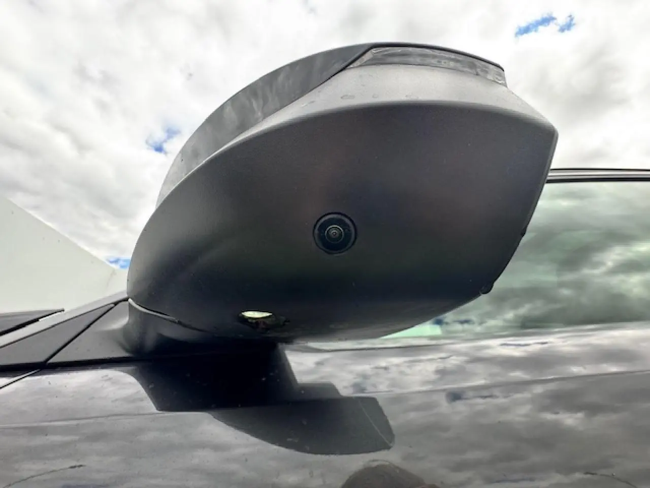 Gros plan sur le rétroviseur noir du côté gauche du Peugeot 5008 gris titane avec caméra intégrée sous un ciel nuageux.