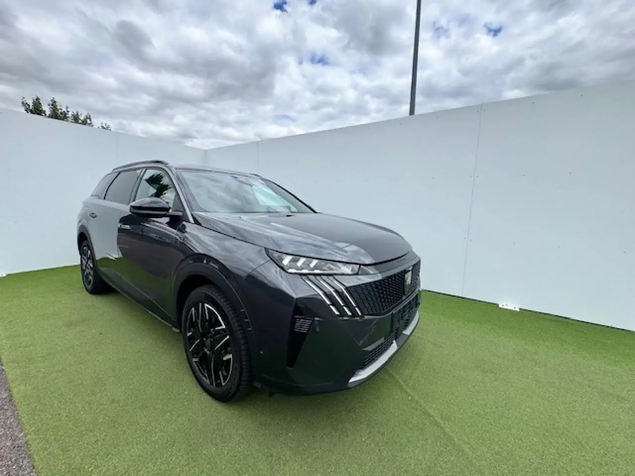 Peugeot 5008 gris titane toit noir en 3/4 avant droit sur fond vert et ciel nuageux.