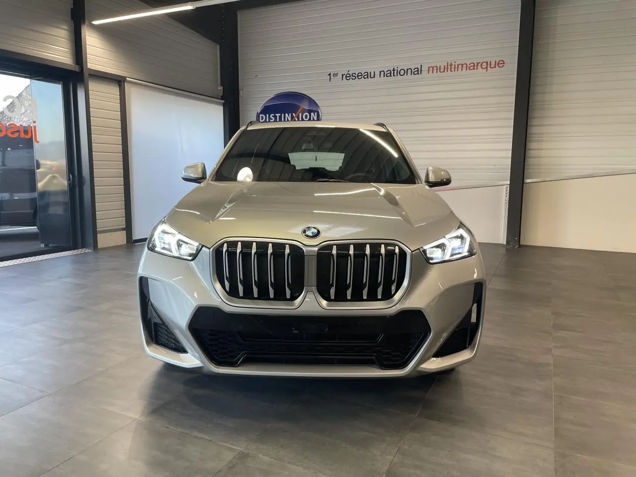 Vue frontale d'un BMW X1 xDrive 20d 2024 en couleur Spacesilber Metallic avec calandre M Sport et phares LED allumés.