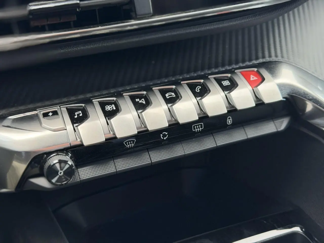 Gros plan sur la rangée de commandes argentées du tableau de bord du Peugeot 3008 noir, boutons fonctionnels et alerte rouge.