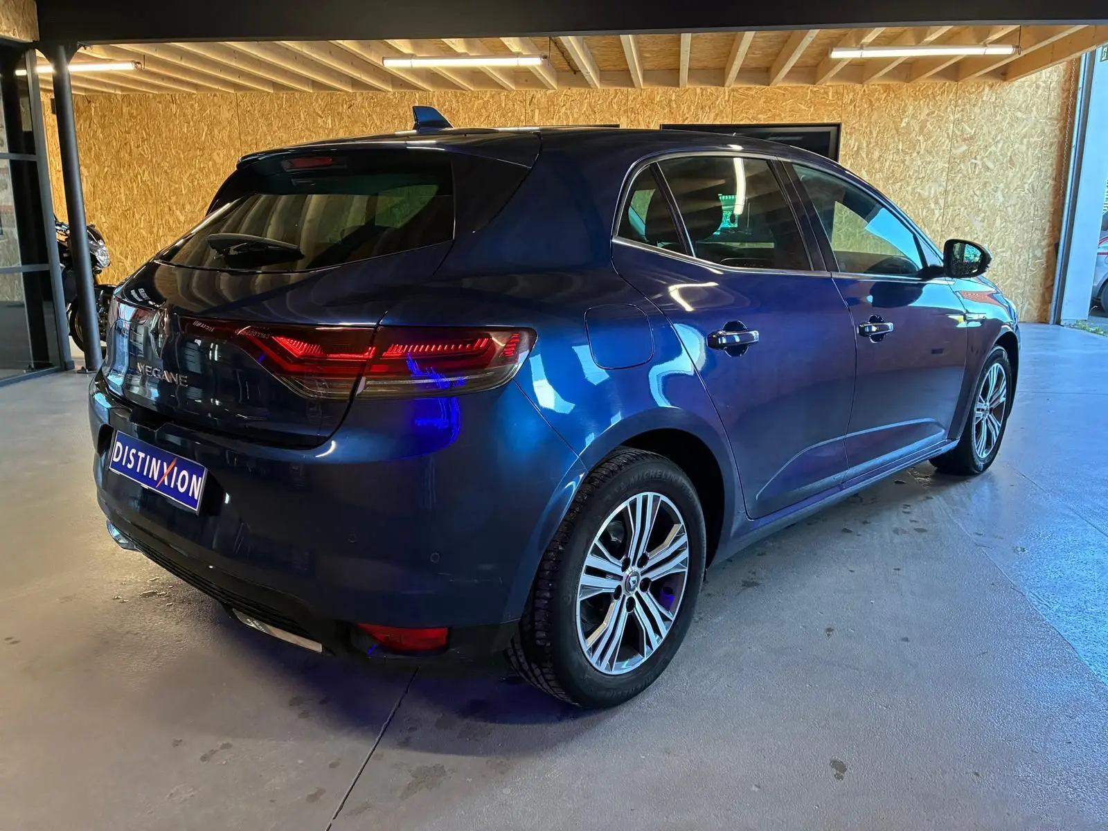 Renault Mégane bleu 3/4 arrière droit en intérieur, feux arrière allumés et jantes alliage biton visibles.