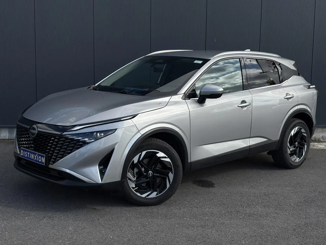 Nissan Qashqai gris argent en 3/4 avant droit, avec jantes alliage noires et toit avec barres chromées.