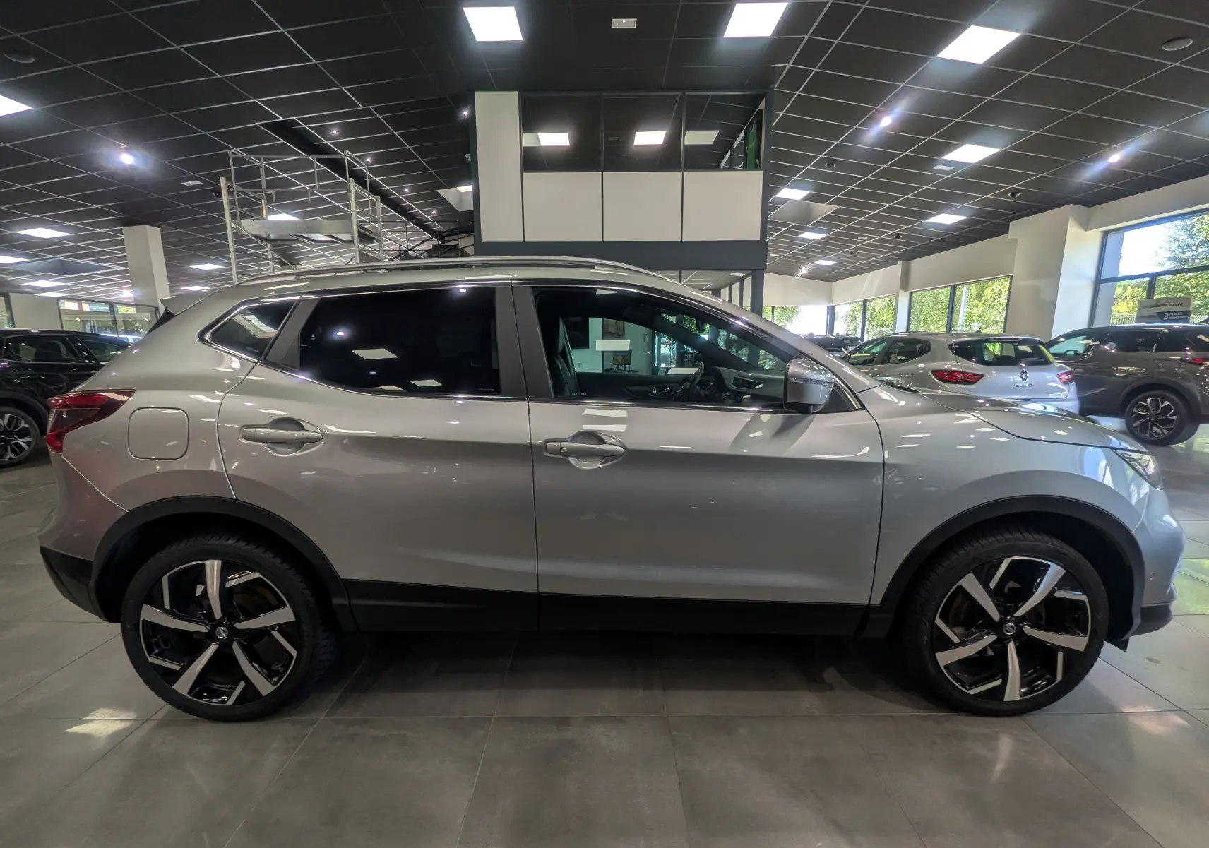 Vue latérale droite d'un Nissan Qashqai gris perle en showroom, avec jantes alliage bicolores et vitres teintées.