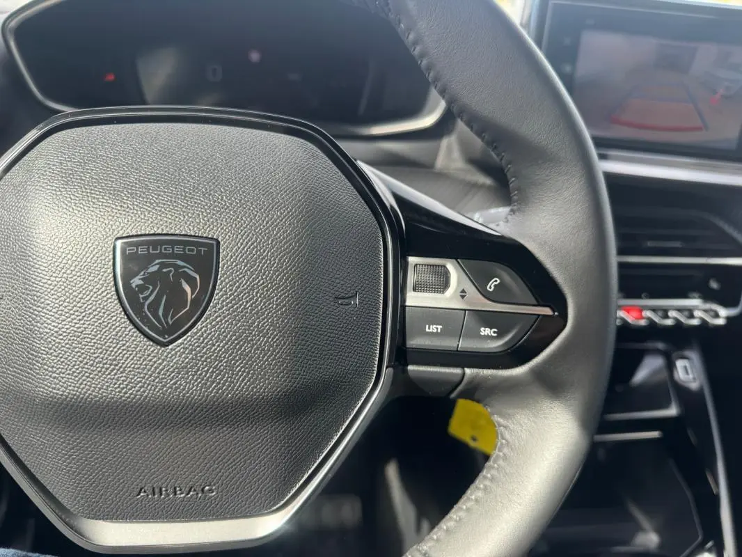 Vue rapprochée du volant noir du Peugeot 2008 2024 avec commandes intégrées et écran tactile en arrière-plan.