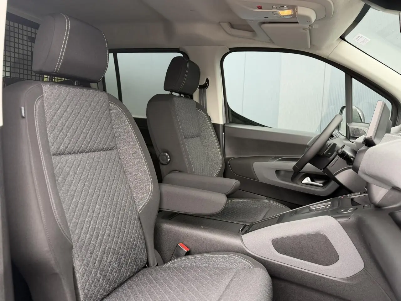 Vue intérieure côté conducteur du Peugeot Rifter 2026, sièges tissu gris avec accoudoir central et tableau de bord moderne.