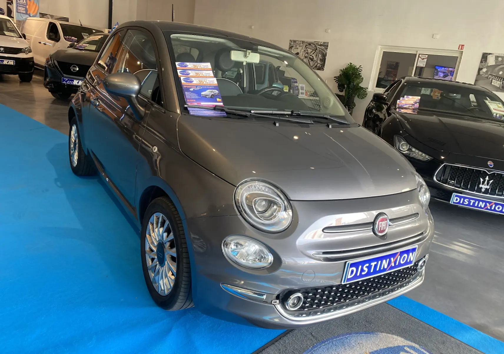 Vue 3/4 avant d'une FIAT 500 gris foncé en showroom, avec jantes multibranches et plaque Distinxion visible.