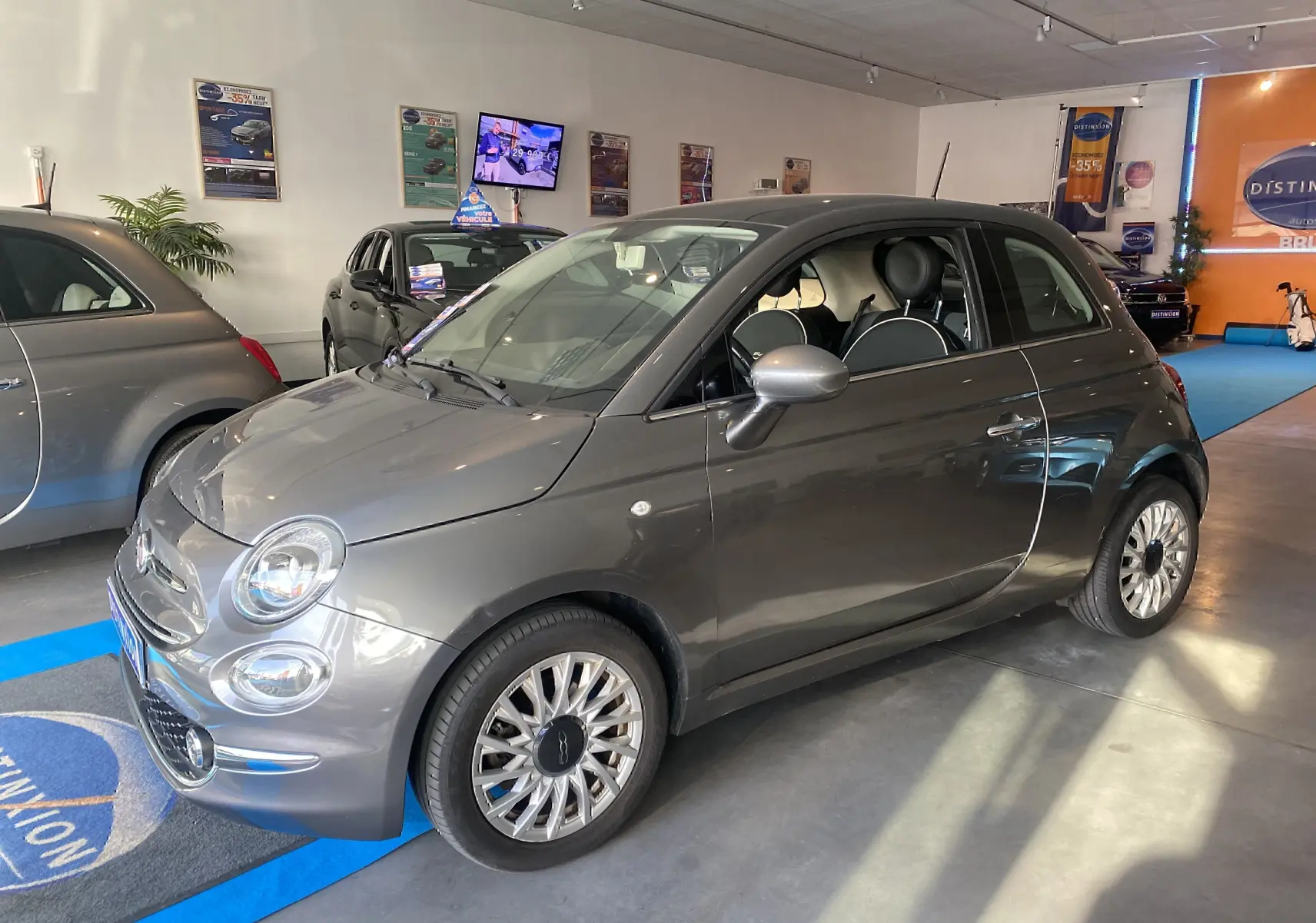 Fiat 500 gris foncé vue 3/4 avant droit en showroom avec jantes multibranches et intérieur noir.