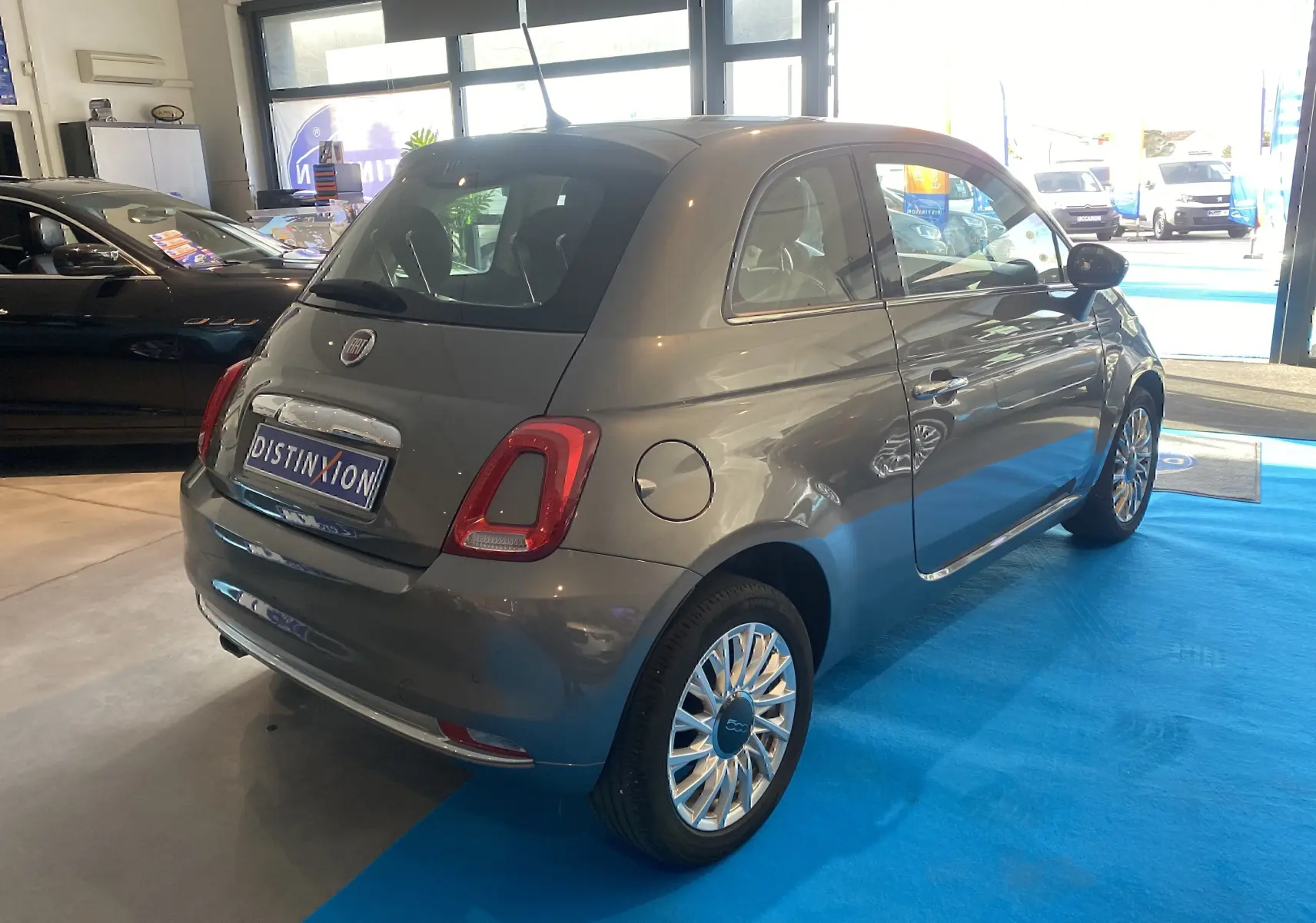 Vue 3/4 arrière droite d'une Fiat 500 gris foncé exposée en showroom avec jantes multibranches chromées.