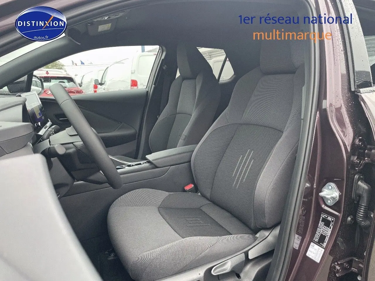 Vue intérieure côté conducteur du Toyota C-HR 1.8 Hybrid 2024 en améthyste métal, sièges tissu gris et console centrale visible.