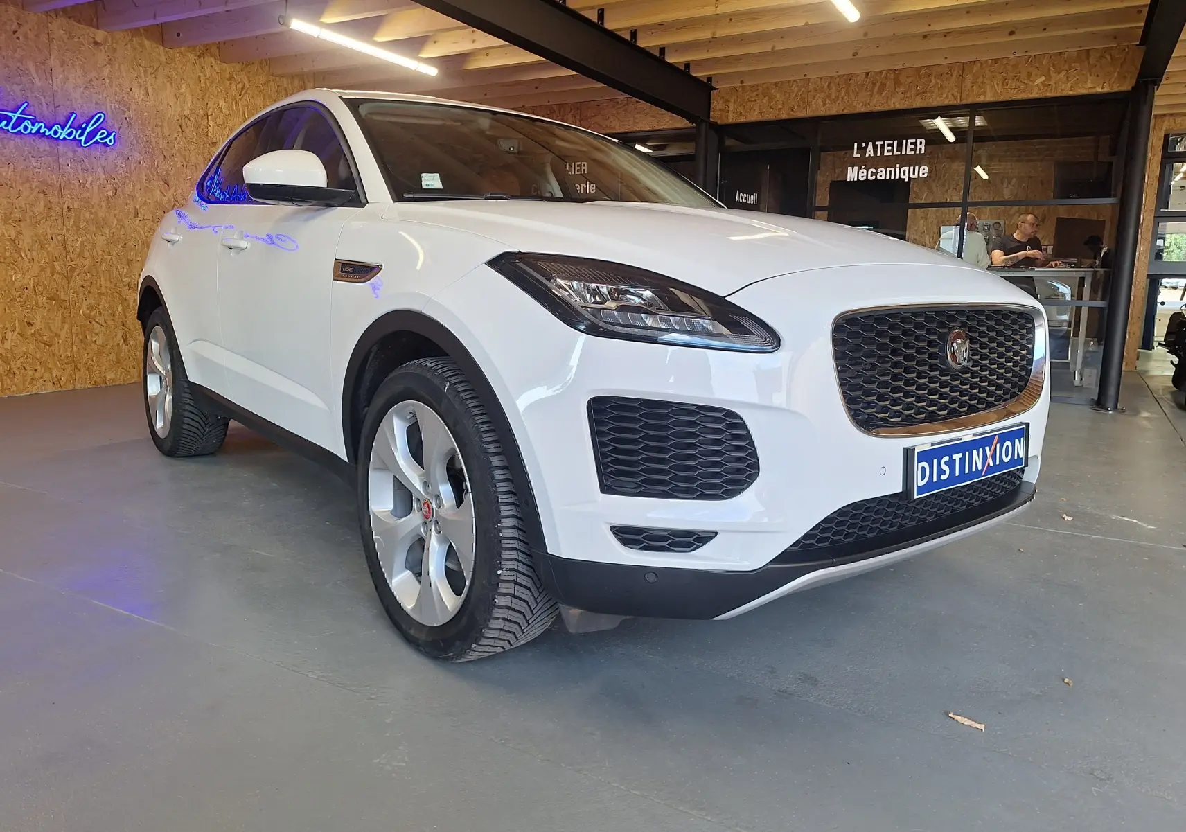Jaguar E-PACE blanc vue 3/4 avant droit en intérieur, avec calandre noire et phares LED allumés.