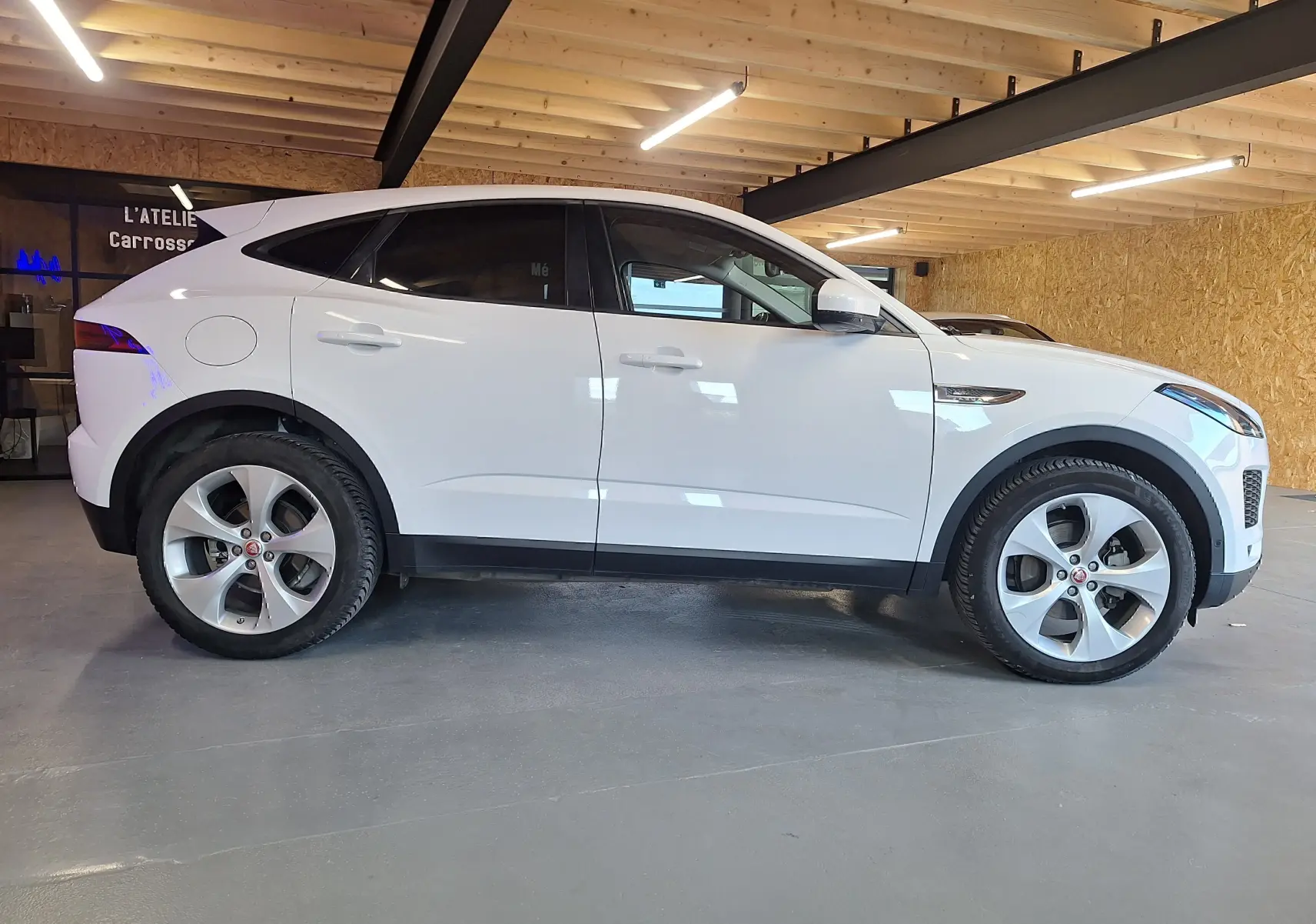 Profil côté gauche d'un Jaguar E-PACE blanc 2019 dans un garage avec toit en bois et sol gris.