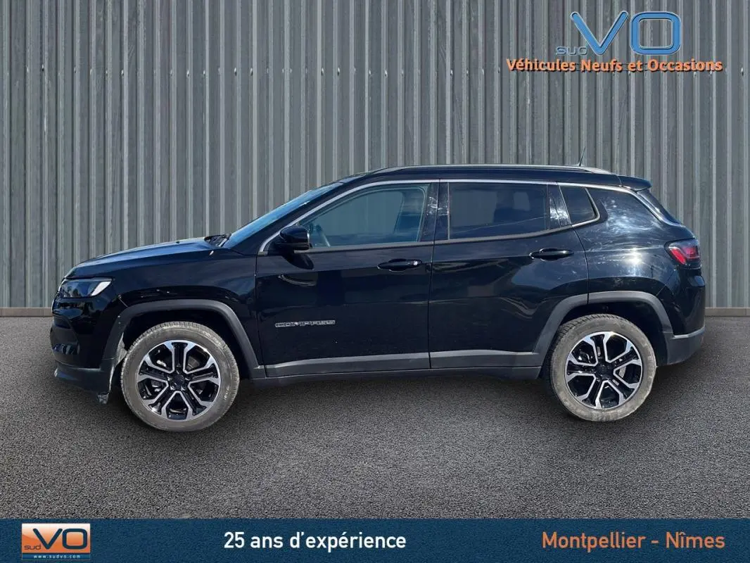 Profil latéral gauche d'un Jeep Compass noir 2023 avec jantes 18 pouces et barres de toit visibles sur fond industriel.