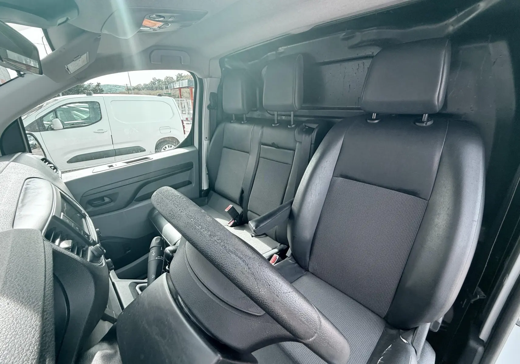 Vue intérieure côté conducteur du Citroën Jumpy blanc, montrant les sièges noirs en cuir et tissu ainsi que le volant.