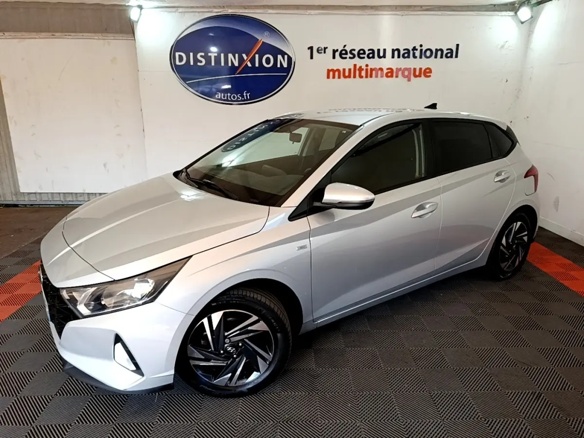 Vue 3/4 avant droite d'une Hyundai i20 gris clair 2022 avec jantes noires et vitres teintées en intérieur showroom.