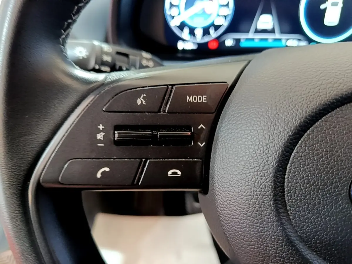 Gros plan sur les commandes multifonctions en cuir du volant gauche de la Hyundai i20 gris clair, tableau de bord numérique en arrière-plan.