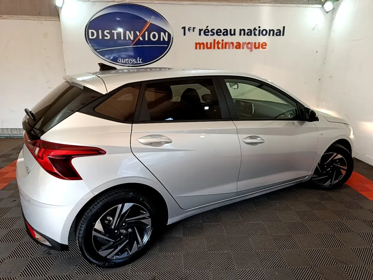 Vue côté droit arrière d'une Hyundai i20 gris clair 2022 avec jantes noires et toit noir contrasté en intérieur showroom.