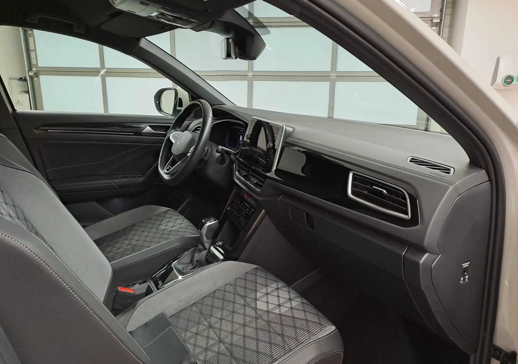 Intérieur du Volkswagen T-ROC 2024 vu côté conducteur, sièges tissu gris à motif losanges et tableau de bord noir brillant.
