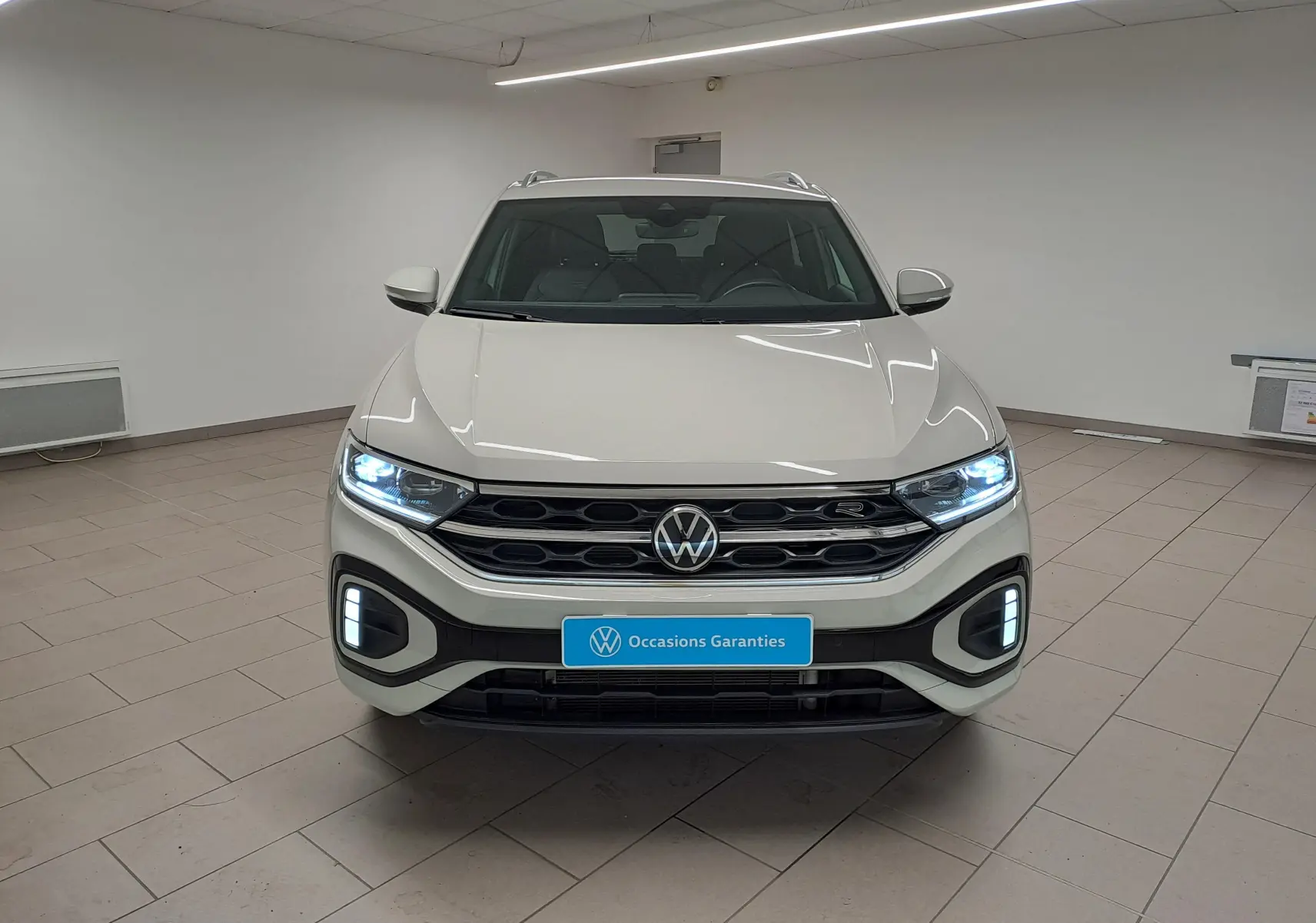 Vue frontale du Volkswagen T-Roc 1.5 TSI R-Line 2024 en Gris Ascot, avec phares LED allumés dans un showroom.
