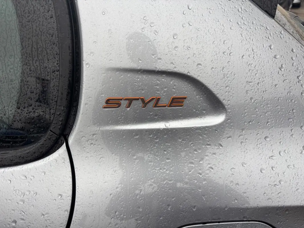 Gros plan sur l’inscription "STYLE" orange sur la portière gris clair d’une Peugeot 208 Hybrid 2025, avec gouttes de pluie.
