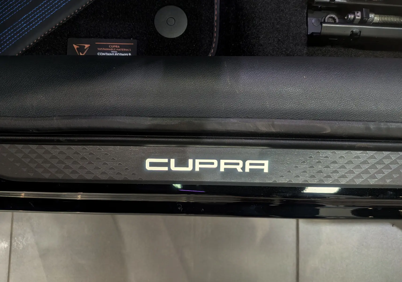 Gros plan sur la plaque de seuil de porte avant avec le logo CUPRA lumineux, intérieur noir et surpiqûres bleues visibles.