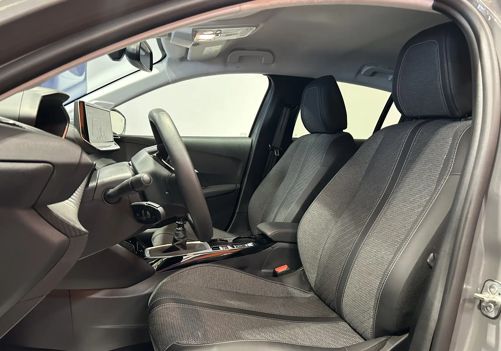 Vue intérieure côté conducteur de la Peugeot 208 gris Selenium, montrant les sièges tissu et la console centrale avec levier de vitesses manuel.