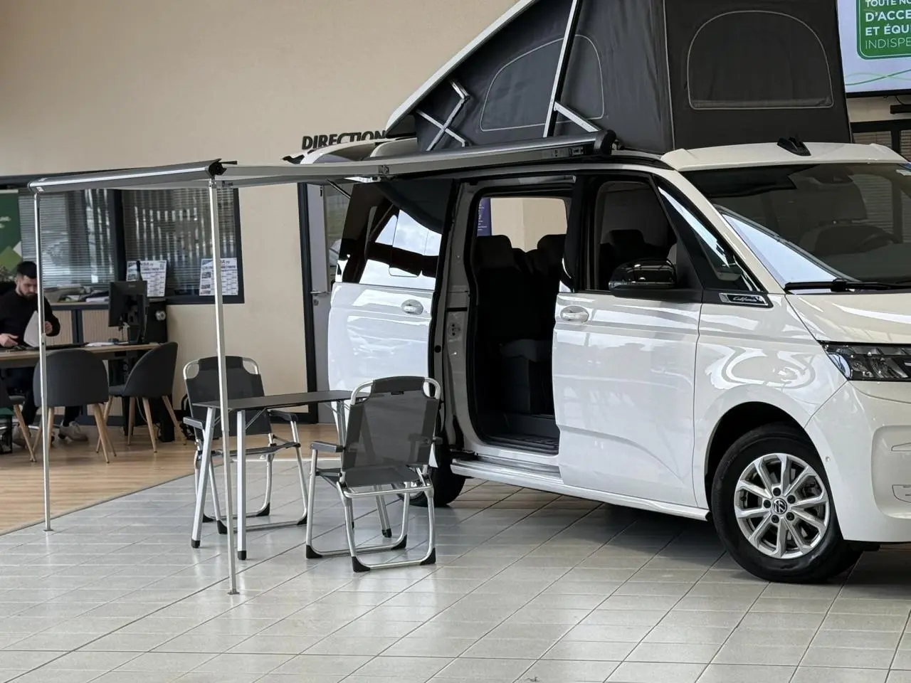 Vue de côté droit d’un Volkswagen California T7 blanc Candy avec toit relevable noir et auvent déployé, porte coulissante ouverte.
