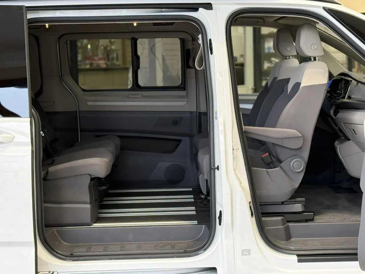 Vue latérale droite intérieure du Volkswagen California T7 blanc Candy montrant les sièges avant avec accoudoirs et la banquette arrière.