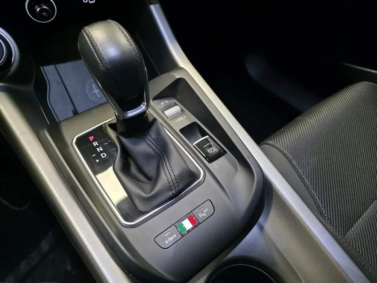 Gros plan sur la console centrale de l'Alfa Romeo Tonale gris, avec levier de vitesse automatique et bouton aux couleurs italiennes.