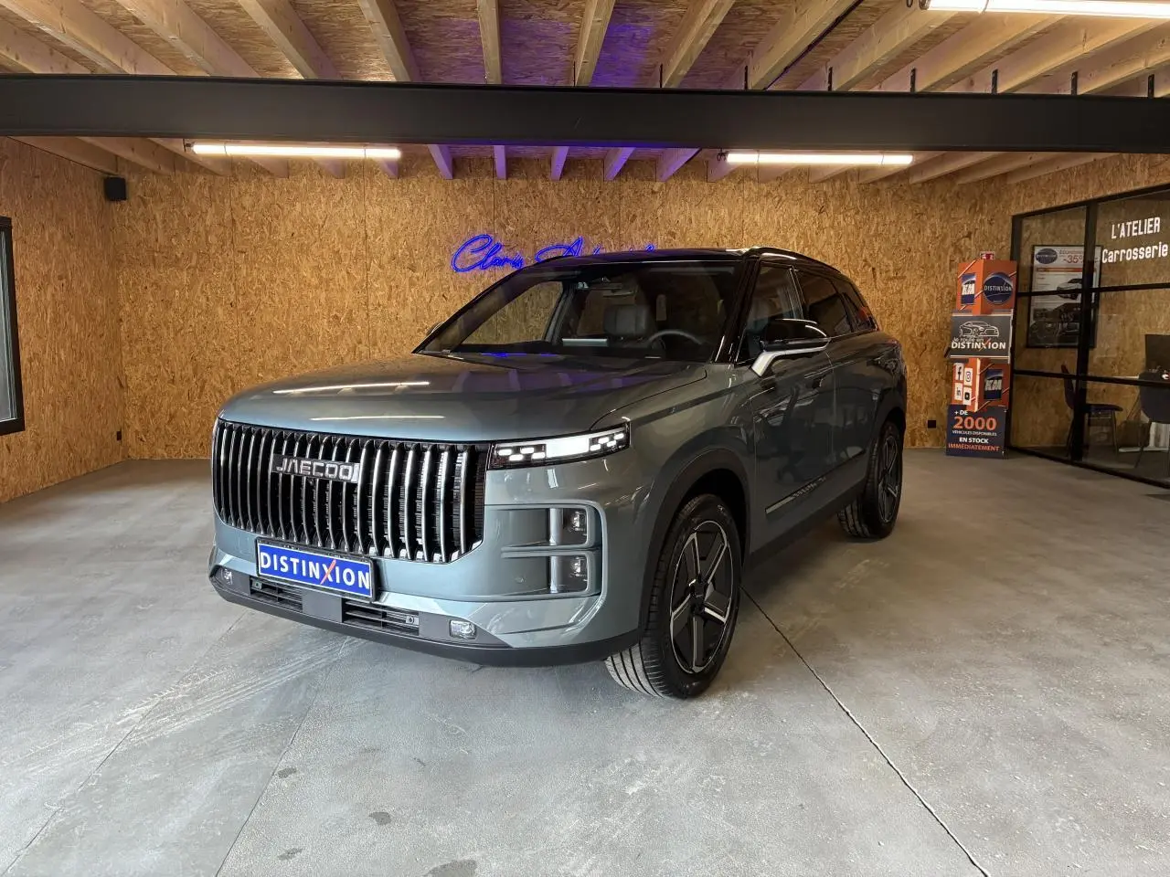 SUV JAECOO J7 gris olive en 3/4 avant droit, avec calandre verticale noire et toit noir dans un garage.