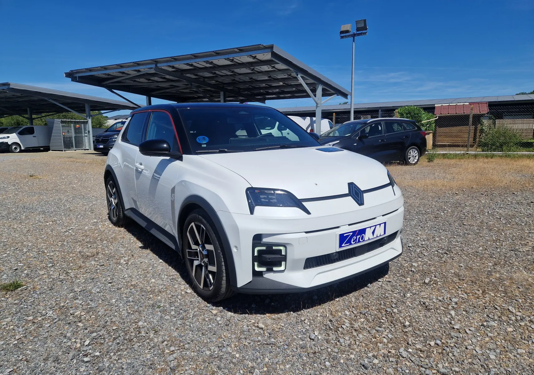 Renault R5 E-TECH blanc nacré et noir en 3/4 avant droit sur parking avec plusieurs fourgons blancs en arrière-plan.