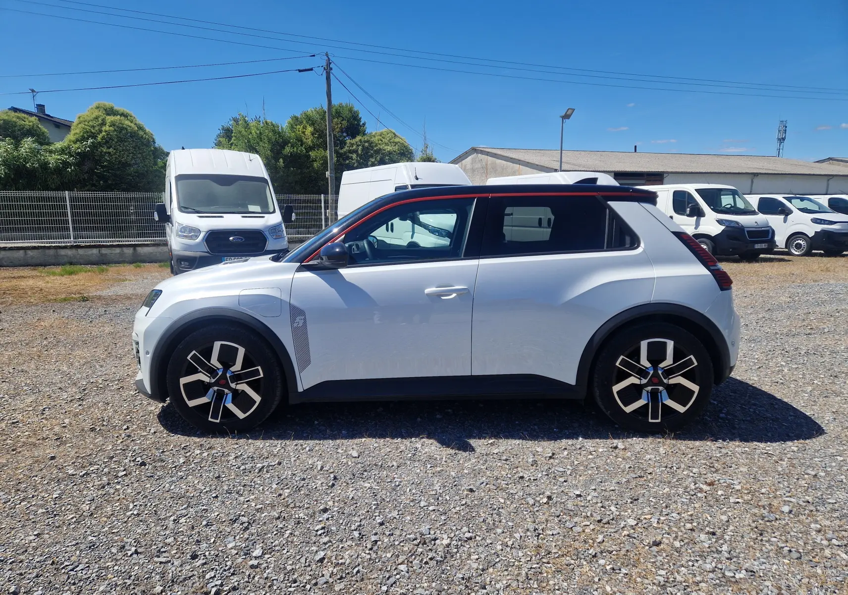 Profil droit de la Renault R5 E-TECH 2025 blanc nacré avec toit noir et jantes stylisées sur un parking extérieur.