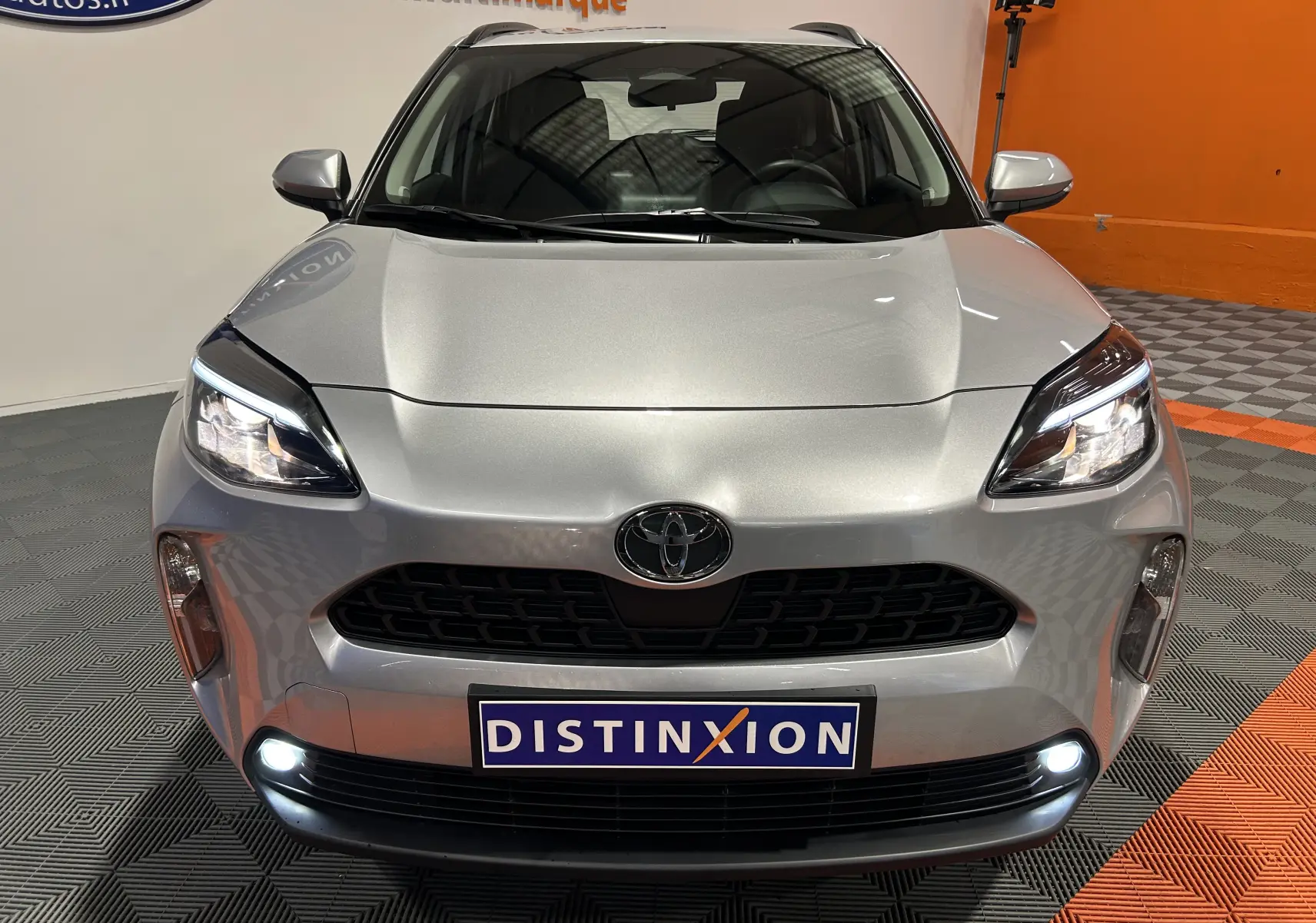 Vue frontale d'une Toyota Yaris Cross gris minéral avec phares et feux de jour allumés dans un showroom.