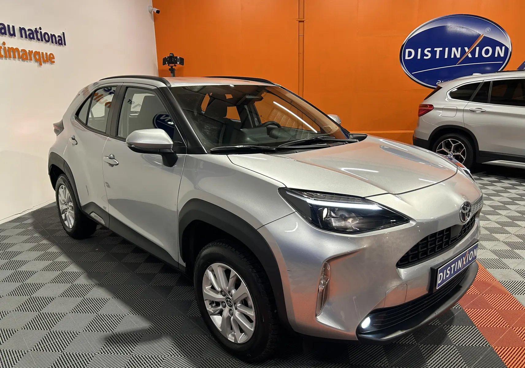 Toyota Yaris Cross gris minéral vue 3/4 avant droit en intérieur avec phares et antibrouillards allumés.