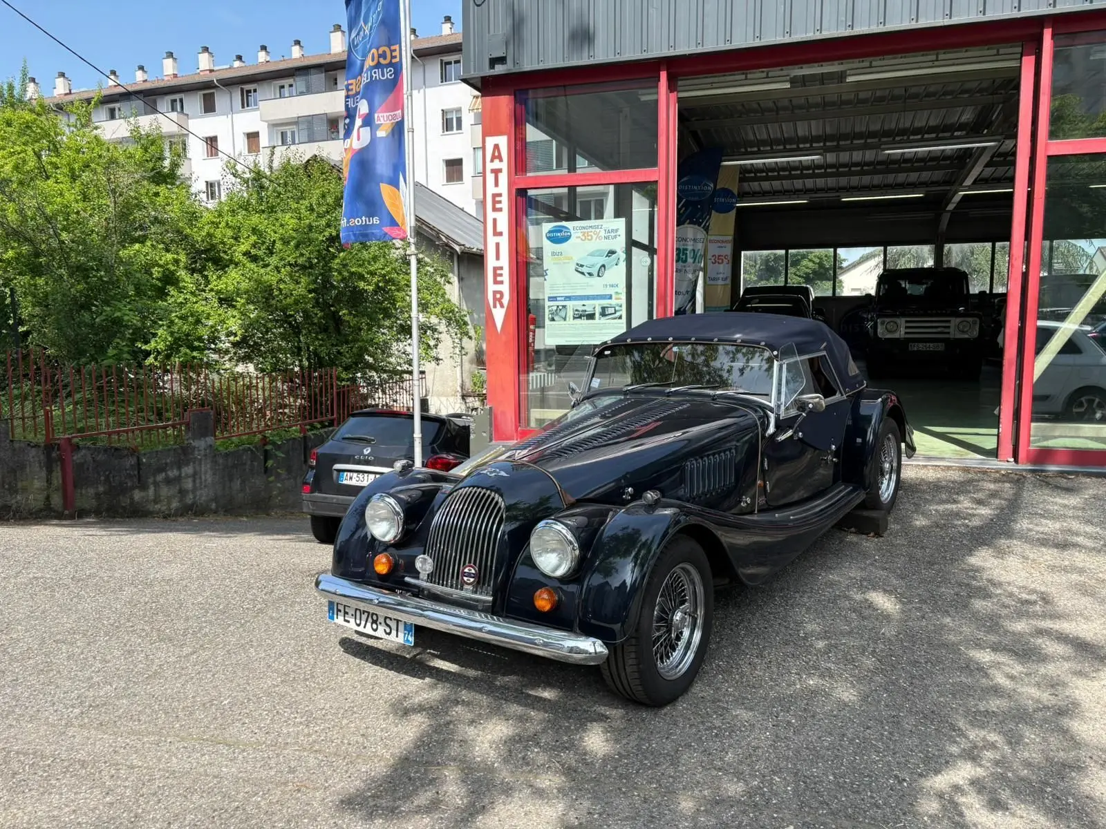 Morgan Plus 4 bleu foncé vue 3/4 avant droit, capote noire et calandre verticale chromée devant un atelier.