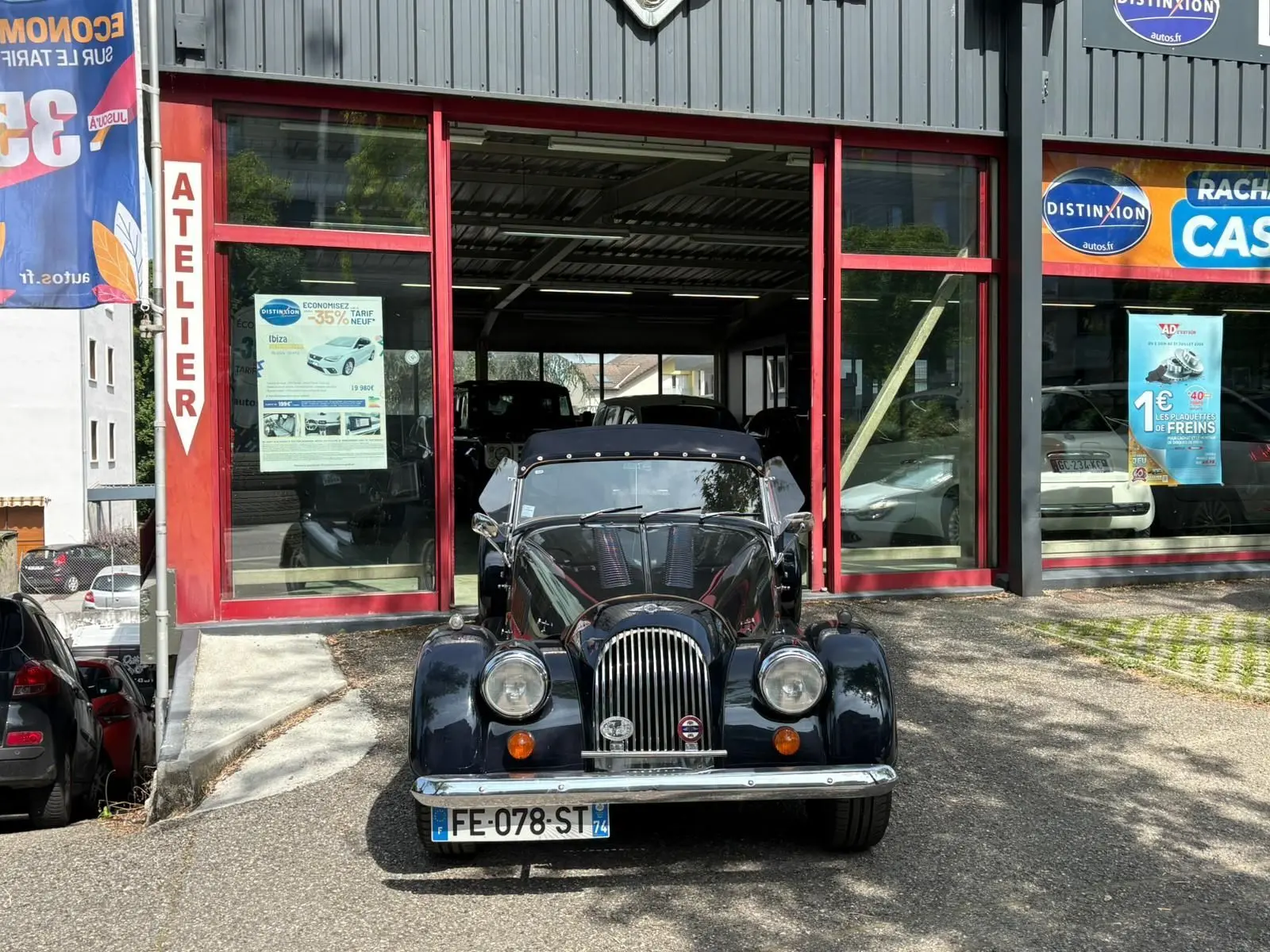 Morgan Plus 4 bleu foncé vue de face, capote noire et calandre chromée devant un atelier automobile.