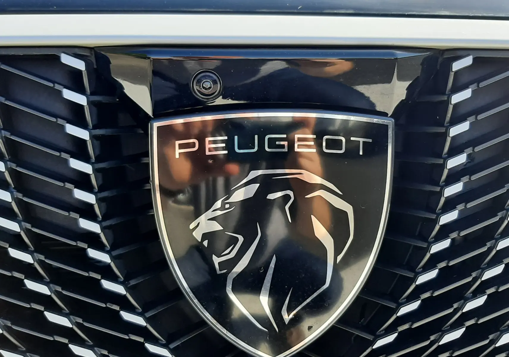 Gros plan sur la calandre noire et le logo Peugeot lion argenté du 3008 blanc Okenite, avec caméra intégrée au-dessus.