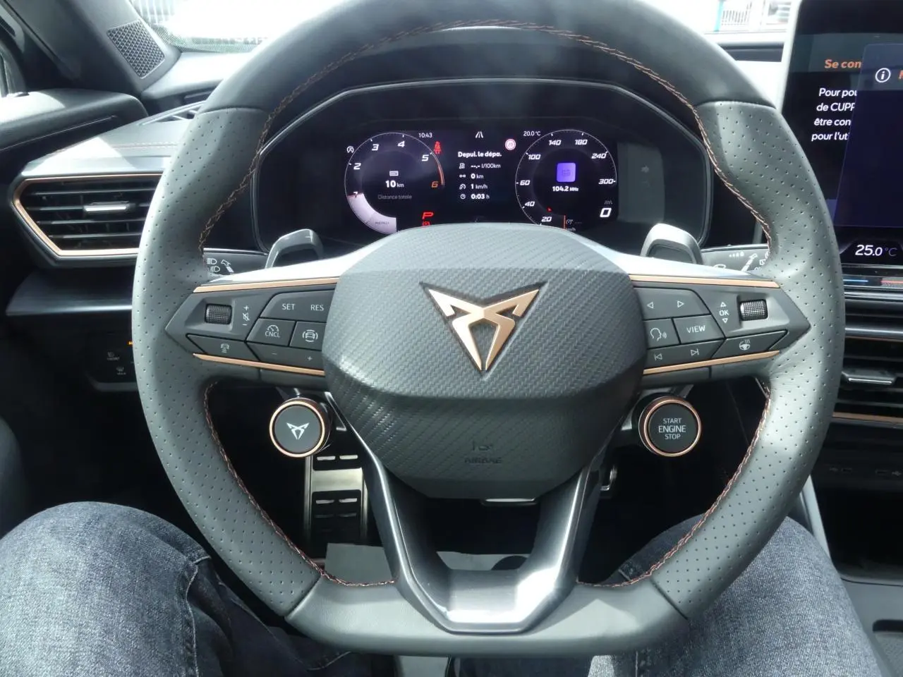 Vue rapprochée du volant perforé noir avec surpiqûres cuivrées et logo CUPRA sur tableau de bord digital.