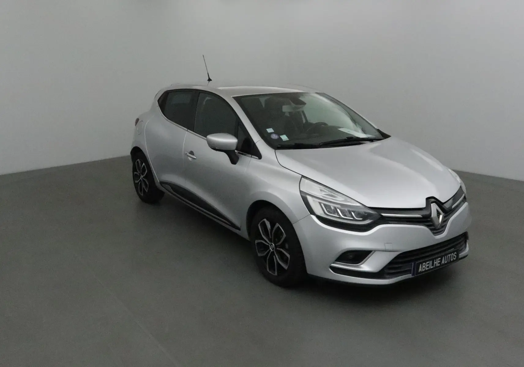 Renault Clio IV gris clair métal vue 3/4 avant droit, avec jantes biton et calandre distinctive Renault.